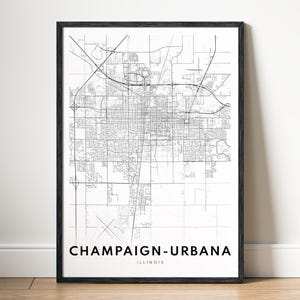 May include: A framed black and white map print of Champaign-Urbana, Illinois. The detailed map shows the city's road network and urban layout. The city name is printed in bold, black text at the bottom of the print.