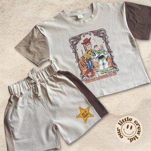 Puede incluir: Conjunto de camiseta y pantalón corto beige. La camiseta tiene mangas marrones y un gráfico de Woody y Buzz Lightyear con el texto "YOU GOT A FRIEND IN ME". Los pantalones cortos tienen una raya marrón y una insignia de sheriff amarilla.