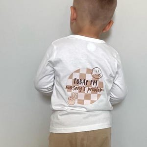 May include: White long-sleeved top with a graphic on the back. The graphic says "TODAY I'M nursery's problem" in brown text, with a smiley face and a peace sign.