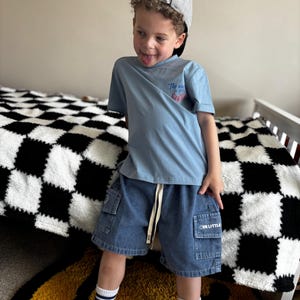 May include: A light blue t-shirt with the text "The world is your oyster" and denim shorts with the text "OUR LITTLE". The shorts have pockets and a drawstring. The person is wearing a gray baseball cap and white socks with blue stripes.