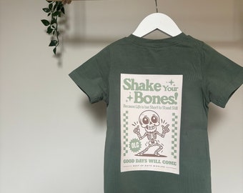 May include: A sage green short-sleeve t-shirt featuring a rectangular graphic on the back. The print displays a cartoon skeleton with the text "Shake Your Bones! Because Life is too Short to Stand Still" and "GOOD DAYS WILL COME" in white and green.