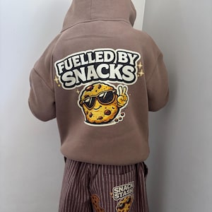 Pode incluir: Camisola com capuz castanha com o texto "FUELLED BY SNACKS" e um biscoito de desenho animado usando óculos de sol. Calças listradas castanhas combinando com um patch "SNACK STASH" e gráfico de biscoito. Roupas casuais infantis.
