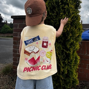 Op de afbeelding: Een lichtgeel mouwloos shirt met een grafische print van een sandwich, koekje, jampot, donut, appelsapverpakking en watermeloenpartjes met de tekst "PICNIC CLUB" en een bruine baseballpet met een ronde patch op de voorkant.