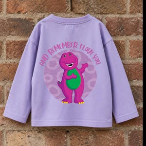 Könnte beinhalten: Ein langärmeliges lavendelfarbenes Shirt mit einer Grafik des violetten Dinosauriers Barney auf der Vorderseite. Das Design zeigt einen kreisförmigen violetten Hintergrund mit Ringmuster und den Text "AND REMEMBER I LOVE YOU" darüber.