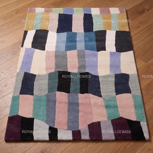 Könnte beinhalten: Ein rechteckiger Teppich mit einem farbenfrohen Patchwork-Design. Der Teppich hat vertikale und horizontale Blöcke in Blau-, Rosa-, Grün-, Grau- und Schwarztönen. Der Text "ROYALLOOMSS" ist auf dem Teppich aufgedruckt.