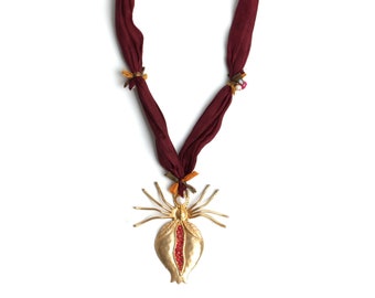 May include: A unique necklace featuring a burgundy fabric cord adorned with small decorative accents. The pendant is a gold-toned, stylized pomegranate with a red interior. The necklace is displayed against a white background.