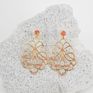Può includere: Un paio di orecchini in filigrana color oro con accenti color corallo. Gli orecchini presentano un delicato design traforato con un motivo floreale o a forma di foglia. Gli orecchini sono esposti su una superficie strutturata grigio chiaro.