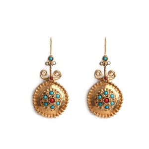 Może przedstawiać: Para złotych kolczyków o okrągłym wzorze. Każdy kolczyk ma centralny czerwony kamień otoczony turkusowymi kamieniami. Kolczyki mają ozdobną górę z turkusowym kamieniem i haczyk do ucha.