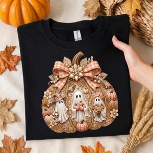 Peut inclure: Un t-shirt noir orné d'un grand motif complexe représentant une citrouille décorée d'un nœud à carreaux, de perles et de fleurs. À l'intérieur de la citrouille se trouvent trois fantômes blancs, dont un portant un chapeau et des fleurs.