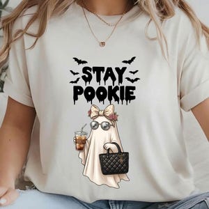 Peut inclure: Un t-shirt couleur crème avec un motif représentant un fantôme portant un nœud à carreaux, des fleurs et des lunettes de soleil boule à facettes. Le fantôme tient un café glacé et un sac à main noir matelassé. Le texte STAY POOKIE est inscrit en noir.