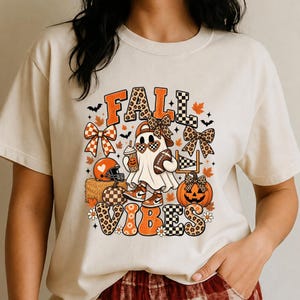 Peut inclure: Un t-shirt couleur crème avec une impression graphique portant le texte FALL VIBES. Le motif représente un fantôme portant des lunettes de soleil et une casquette, tenant un ballon de football et une boisson, entouré de citrouilles, de nœuds, de feuilles d'automne et de chauves-souris.