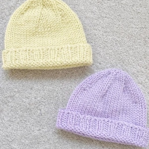 May include: Two knitted beanies. One is a pale yellow color, and the other is a light purple color. Both beanies have a folded brim and are made of a thick, textured yarn. The beanies are displayed on a gray surface.