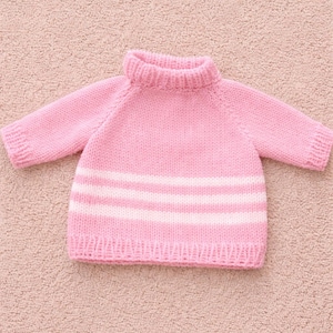May include: A pale pink knitted jumper with a ribbed roll neck and long sleeves. The jumper has three horizontal white stripes across the lower section. The garment is displayed on a textured, pale pink surface.