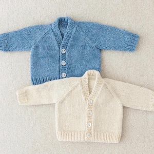 Pode incluir: Dois casaquinhos de bebé de malha, um azul claro e outro creme. Ambos têm decote em V, mangas compridas e fechos de botões. Os casaquinhos são feitos de um fio macio e texturizado, ideais para um recém-nascido.