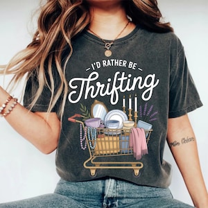 Peut inclure: T-shirt gris foncé avec l'inscription "I'd Rather Be Thrifting" au-dessus d'un dessin de chariot de magasinage rempli d'articles d'occasion. Le chariot comprend un miroir, de la vaisselle, des bougies et une écharpe rose. Un collier et d'autres articles sont également visibles.
