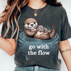 May include: Dark grey t-shirt with an otter illustration. A larger otter holds a smaller one in water. The text "go with the flow" is below the otters in white.