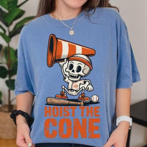 May include: A light blue t-shirt featuring a cartoon skeleton in a baseball uniform holding an orange and white traffic cone. The text "HOIST THE CONE" is printed in orange below the skeleton.