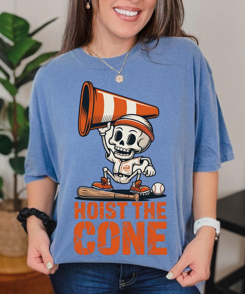May include: A light blue t-shirt featuring a cartoon skeleton in a baseball uniform holding an orange and white traffic cone. The text "HOIST THE CONE" is printed in orange below the skeleton.