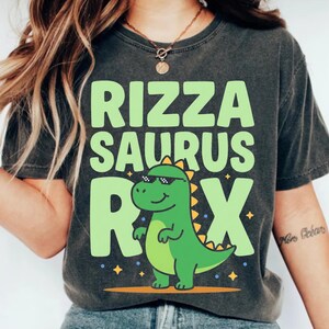 Puede incluir: Camiseta gris oscuro con las palabras "RIZZA SAURUS REX" en verde claro, con un dinosaurio de dibujos animados con gafas de sol. El dinosaurio es verde con detalles amarillos y está sobre una línea amarilla.