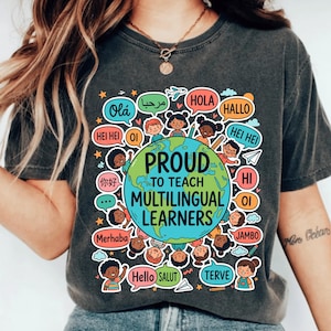 May include: A charcoal gray t-shirt featuring a colorful graphic of a globe surrounded by diverse children and speech bubbles containing various greetings. The central text reads PROUD TO TEACH MULTILINGUAL LEARNERS in bold, dark blue capital letters.