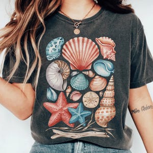 May include: A charcoal gray t-shirt featuring a graphic print of various seashells, including a large pink scallop, a nautilus, a sand dollar, a red starfish, a blue starfish, and a tall spiral shell, arranged above a piece of driftwood.