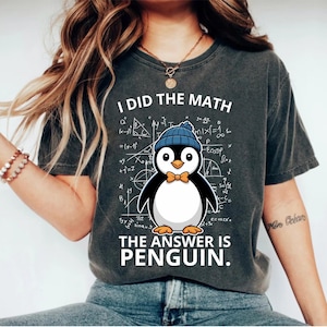 Op de afbeelding: Donkergrijs t-shirt met de tekst "I DID THE MATH THE ANSWER IS PENGUIN." Een cartoon pinguïn met een blauwe muts en strikdas, omringd door wiskundige formules.