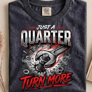 May include: A charcoal gray t-shirt featuring a graphic print of a wrench tightening a bolt into a cracked surface. The text reads "JUST A QUARTER TURN MORE" in bold white and red lettering. The design includes scattered metal nuts and bolts.