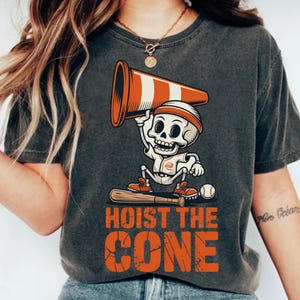 May include: Dark gray t-shirt featuring a cartoon skeleton holding a traffic cone above its head. The skeleton is wearing a baseball uniform and a headband. The text "HOIST THE CONE" is printed in orange below the skeleton.