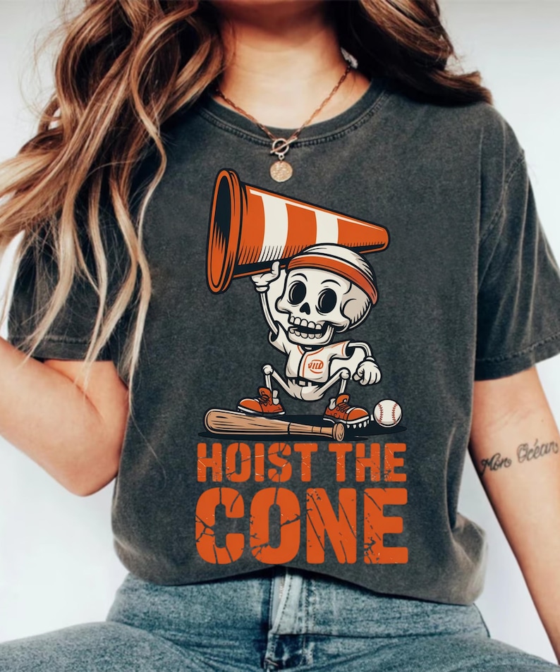 May include: Dark gray t-shirt featuring a cartoon skeleton holding a traffic cone above its head. The skeleton is wearing a baseball uniform and a headband. The text "HOIST THE CONE" is printed in orange below the skeleton.