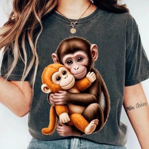 May include: Dark gray t-shirt featuring a graphic of two cartoon monkeys hugging. One monkey is brown, and the other is orange. The monkeys have large eyes and are smiling. The shirt is made of a soft material.