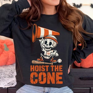 May include: Black long-sleeve shirt featuring a Halloween-themed graphic. The design includes a skeleton holding a traffic cone, a baseball bat, and a baseball. The text "HOIST THE CONE" is printed in orange.