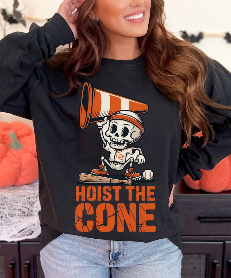 May include: Black long-sleeve shirt featuring a Halloween-themed graphic. The design includes a skeleton holding a traffic cone, a baseball bat, and a baseball. The text "HOIST THE CONE" is printed in orange.