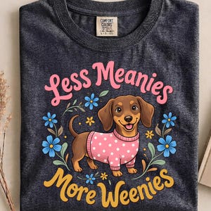 Puede incluir: Una camiseta gris carbón con un perro salchicha marrón que lleva un suéter rosa de lunares, rodeado de flores azules y detalles amarillos. El texto dice "Less Meanies" en rosa y "More Weenies" en amarillo, con una etiqueta Comfort Colors.