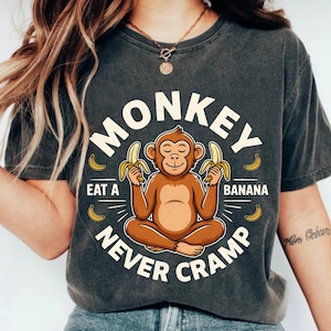以下が含まれることがあります： チャコールグレーの半袖Tシャツ。蓮華座で座り、2本のバナナを持っている茶色の猿のグラフィックが描かれています。クリーム色のフォントで「MONKEY EAT A BANANA NEVER CRAMP」という文字があり、その周りに小さな黄色いバナナのアイコンが配置されています。