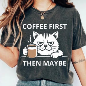 May include: Dark gray t-shirt with the text "COFFEE FIRST THEN MAYBE" and a white illustration of a grumpy cat holding a coffee cup. The cat has a stern expression. The shirt is a casual wear item.