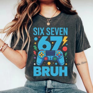 May include: Dark gray t-shirt with a blue graphic that reads "SIX SEVEN 67 BRUH". The graphic includes a blue video game controller, lightning bolts, and colorful dots. The shirt is a casual style.