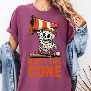 May include: A maroon t-shirt featuring a cartoon skeleton in a baseball uniform holding a traffic cone. The text "HOIST THE CONE" is printed in orange below the skeleton. A baseball bat and ball are also visible.