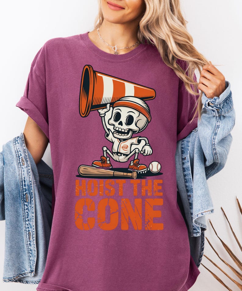 May include: A maroon t-shirt featuring a cartoon skeleton in a baseball uniform holding a traffic cone. The text "HOIST THE CONE" is printed in orange below the skeleton. A baseball bat and ball are also visible.