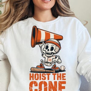 May include: White sweatshirt featuring a cartoon skeleton in a baseball uniform holding a traffic cone. The skeleton is standing on a baseball bat, with a baseball nearby. The text "HOIST THE CONE" is printed in orange.