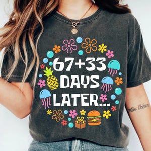 May include: Dark gray t-shirt with a colorful graphic. The graphic features the text "67+33 DAYS LATER..." surrounded by cartoon flowers, bubbles, a pineapple, jellyfish, a gift box, and a hamburger.