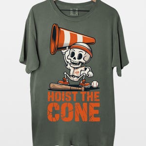 May include: Olive green t-shirt featuring a cartoon skeleton wearing a headband and holding a traffic cone. The skeleton is dressed in a baseball uniform and stands with a bat and ball. The text "HOIST THE CONE" is printed in orange.