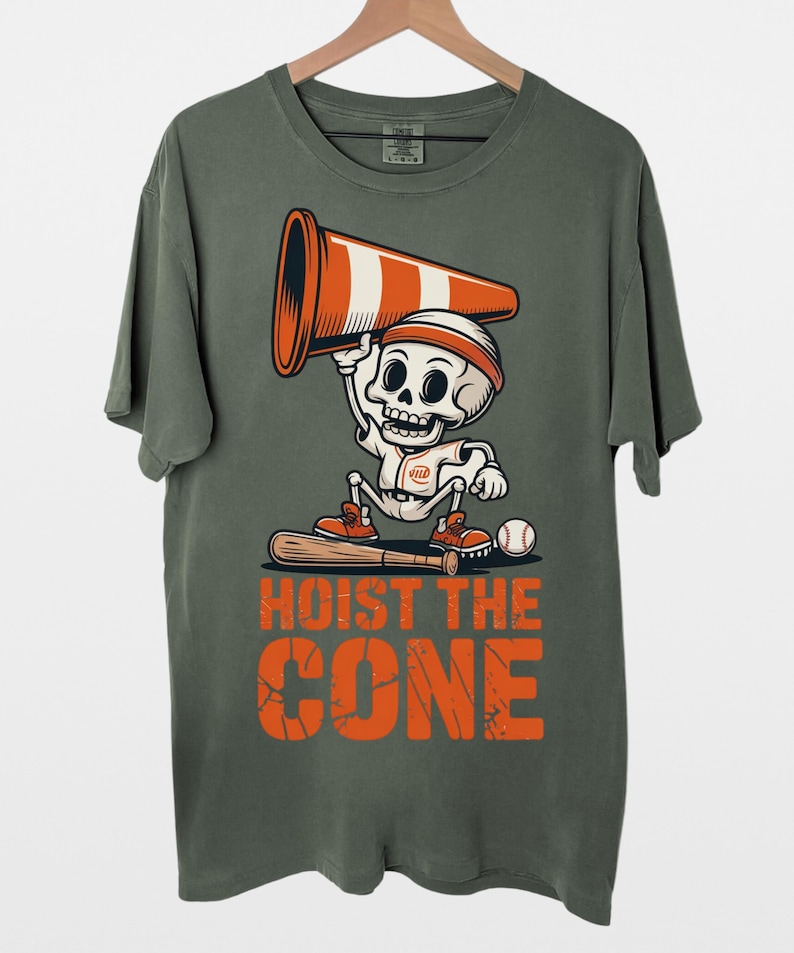May include: Olive green t-shirt featuring a cartoon skeleton wearing a headband and holding a traffic cone. The skeleton is dressed in a baseball uniform and stands with a bat and ball. The text "HOIST THE CONE" is printed in orange.
