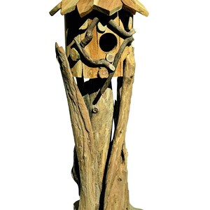 May include: A tall birdhouse constructed from light brown natural wood, featuring a peaked roof, a circular entrance hole, and decorative twig accents. The structure is supported by a thick, vertical wooden post mounted on a flat base.