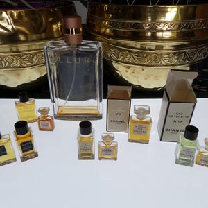 Peut inclure: Une collection de flacons de parfum Chanel comprenant un grand flacon Allure, plusieurs flacons miniatures avec du liquide jaune et transparent, et deux petites boîtes en carton étiquetées No. 5 et No. 19 Eau de Toilette sur fond blanc.