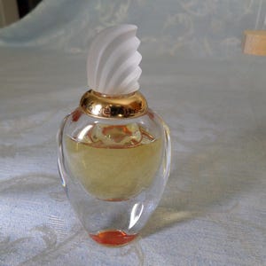 May include: A clear glass bottle of perfume with a gold cap and a white swirl design on the top. The bottle is filled with a light yellow liquid.