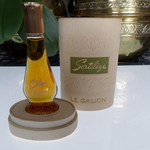 May include: A vintage perfume bottle with a gold-coloured cap and a clear glass body filled with amber liquid. The bottle sits on a round, beige stand. Beside it is a cylindrical container with the text "Sortilège" and "LE GALION".