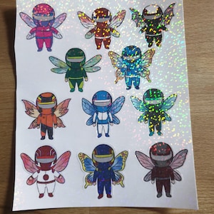 Può includere: Foglio di adesivi con personaggi dei cartoni animati simili a fate, con caschi da corsa e ali. Gli adesivi hanno un effetto olografico e glitterato. Ogni personaggio è di un colore diverso, tra cui rosso, verde, blu e arancione. Gli adesivi sono su sfondo bianco.