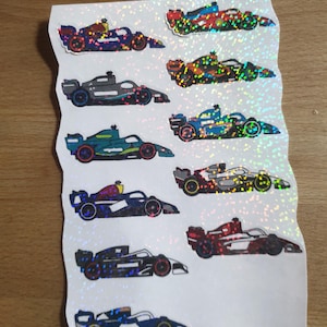 Può includere: Foglio di adesivi olografici con varie auto da corsa di Formula 1 in diversi colori. Gli adesivi presentano disegni dettagliati dei veicoli, con un effetto scintillante e iridescente. Le auto sono in tonalità di blu, rosso, verde e nero.
