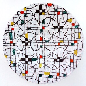 May include: A round ceramic plate with a white background and a black, green, red, and yellow geometric pattern. The pattern includes a grid of lines and circles.