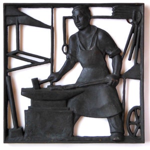 以下が含まれることがあります： 金色の金属製の壁の彫刻で、人が金床で作業をしている様子が描かれている。彫刻は黒い金属製の枠で囲まれている。